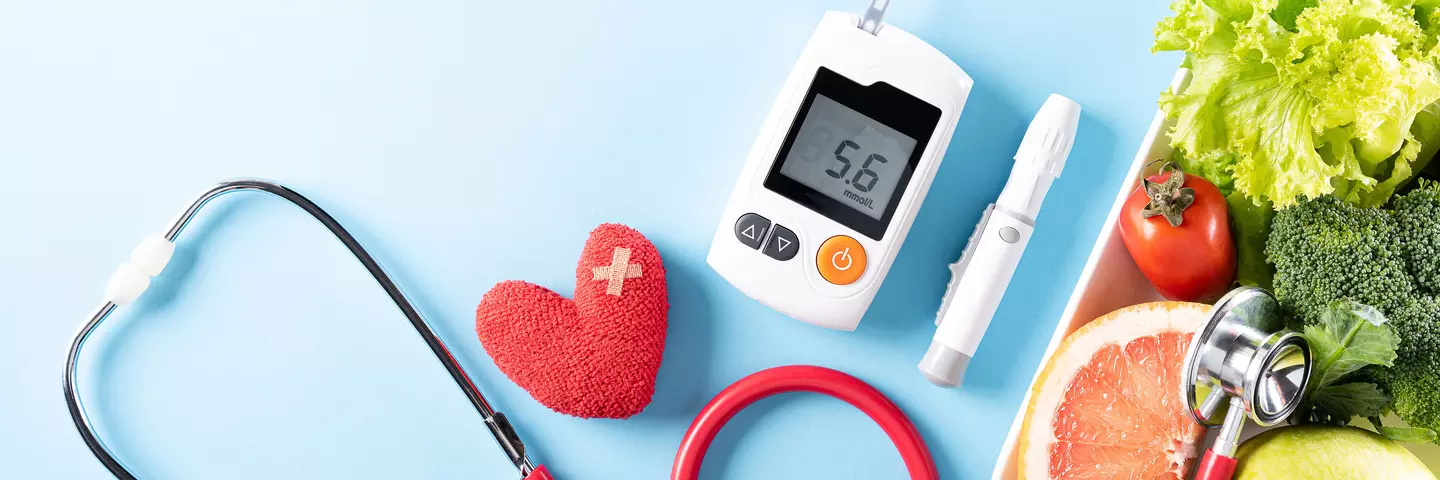 Gesundheits- und Wellnessartikel auf einem blauen Hintergrund, darunter ein rotes Stethoskop, ein herzförmiges Objekt, ein Pflaster, ein Blutzuckermessgerät und frische Lebensmittel wie Salat, Tomate und Grapefruit.
