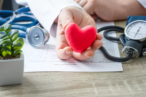 Medizinisches Fachpersonal hält ein rotes Herz mit einem EKG-Symbol in der Hand, im Hintergrund ein Blutdruckmessgerät und eine Pflanze auf einem Schreibtisch.
